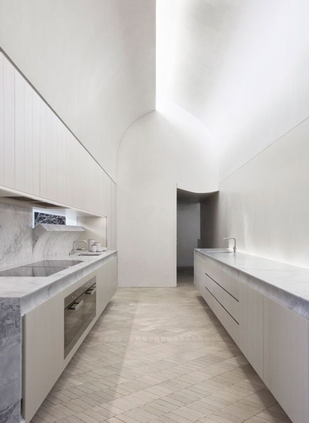 A bright, minimalist galley kitchen with tall white integrated cabinetry on both sides, pale marble or stone benchtops and splashbacks, a large island or peninsula with an inset sink, and a pale herringbone-pattern tiled floor. The space has a high, sculptural ceiling with curved transitions and a dark doorway at the rear. The palette is predominantly white, light grey, and pale stone, creating a clean, refined atmosphere.