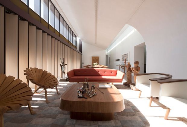 A bright, spacious living room with a sculptural red sofa, rounded timber coffee table, mid-century-style wooden lounge chairs, and a pale upholstered armchair. Full-height vertical timber screening and clerestory glazing line the left side, while a large curved opening and high white ceiling create a strong architectural focal point. Sunlight casts dramatic shadows across a muted patterned rug and warm timber furnishings.