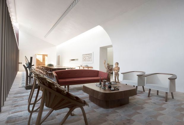 Bright, spacious open-plan living and dining room with a refined mid-century-inspired interior. The room features a long dining table and chairs, a burgundy upholstered sofa, sculptural lounge chairs, a rounded timber coffee table, white built-in walls, an arched opening, and a patterned stone or tile floor. Full-height dark vertical window or wall screening lines one side, while linear ceiling detailing adds architectural interest.