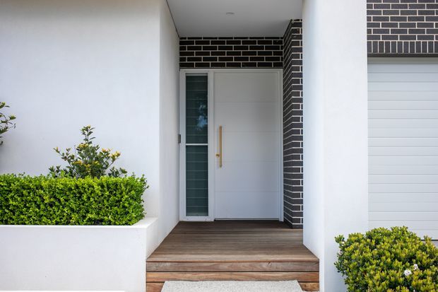 Contemporary residential front entry with a recessed white doorway framed by dark face brick, slim vertical glazing, and a long timber door handle. Timber decking and steps lead to the entrance, with white rendered walls, integrated planter boxes, clipped green shrubs, and a partially visible sectional garage door.