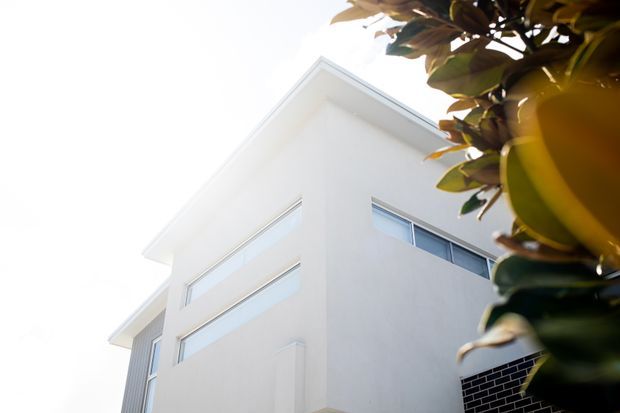 Upward-looking exterior view of a contemporary white multi-storey home, featuring clean rendered walls, a sharply pitched roofline, narrow horizontal windows, and a contrasting section of dark brickwork. Foliage frames the upper-right foreground.