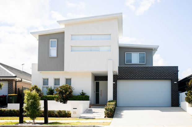 Front exterior view of a contemporary multi-level suburban residence with a white rendered facade, grey vertical cladding, black brickwork around the garage, horizontal highlight windows, and a wide double garage. The home is approached by a paved driveway and landscaped front garden.