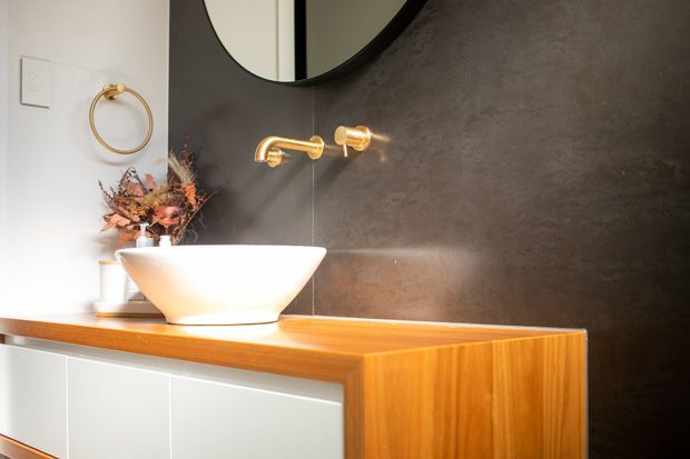 Stylish bathroom vanity featuring a white vessel basin on a warm timber benchtop, white integrated cabinetry, a dark textured wall finish, and brass wall-mounted tapware and circular mirror. A small floral arrangement and brass towel ring add decorative detail.