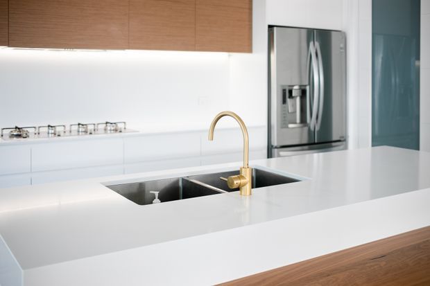Bright contemporary kitchen focused on a large white island with an integrated double-bowl stainless-steel sink and curved brass-gold mixer tap. White cabinetry and splashback create a clean backdrop, while warm timber overhead and island cabinetry add contrast. A stainless-steel refrigerator and blue-tinted glass panel are visible in the background.