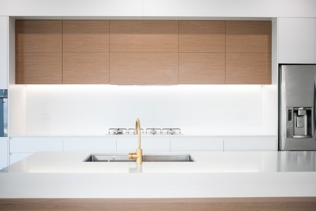 Bright minimalist kitchen focused on a white island with an integrated stainless-steel sink and a brass-toned mixer tap. The rear wall has white cabinetry and a clean white splashback, with extensive light timber overhead cabinetry and a large stainless-steel refrigerator at right. Under-cabinet lighting creates an evenly illuminated, refined interior.