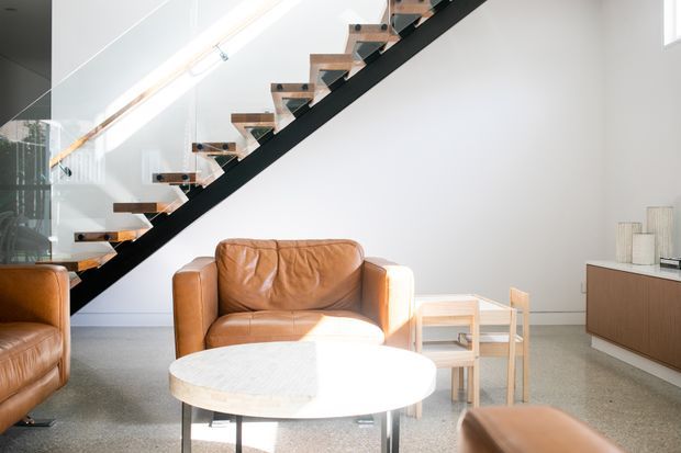 Bright contemporary living room with a sculptural open-riser staircase, timber treads, dark structural stringer, and clear glass balustrade. The space features cognac leather lounge chairs, a round pale stone or terrazzo coffee table, light walls, and a low timber media cabinet.