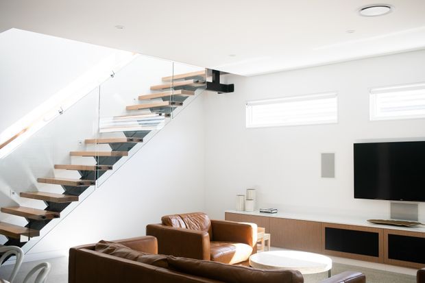 Bright contemporary living room with a floating timber staircase, clear glass balustrade, white walls and ceiling, brown leather seating, and a low timber media console with wall-mounted television. The staircase is supported by a dark central structural element and creates a strong architectural feature.