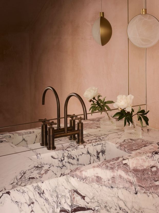 Close-up of a luxurious bathroom vanity featuring a double integrated marble basin with dramatic veining, dark bronze or aged brass curved faucets, a matching marble benchtop and splashback, and warm blush-toned wall panelling. A large mirror reflects the vanity, while sculptural pendant lights and floral arrangements add a refined decorative layer.