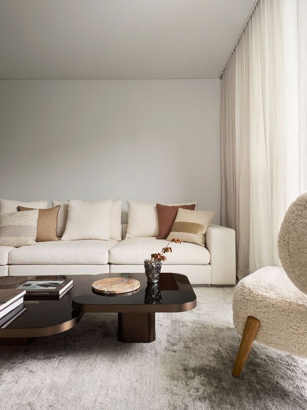 A refined contemporary living room with a low cream sectional sofa, layered neutral cushions, sculptural dark coffee tables, a textured cream accent chair, pale carpet, and full-height sheer curtains. The room is softly lit with a calm neutral palette and subtle warm brown accents.