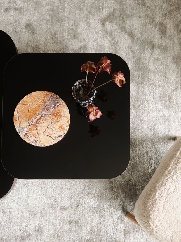Top-down view of a refined living-room vignette featuring a black square coffee table with softly rounded corners, a circular veined stone inlay or tray, and a small dark vase holding pink orchid stems. The table sits on a pale textured rug beside a cream boucle upholstered chair, creating a minimalist, luxe composition with organic natural accents.