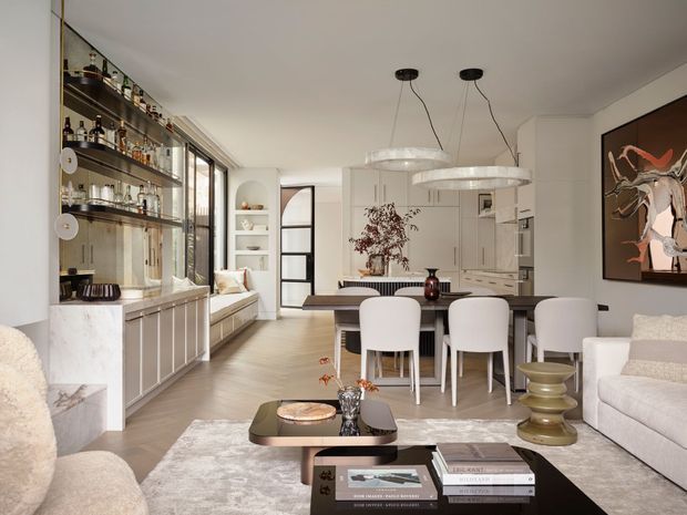 Open-plan contemporary apartment interior combining a living room, dining area, kitchen, and prominent built-in bar display. The space features pale timber flooring, white cabinetry and walls, a marble-clad bar with open shelving, sculptural pendant lighting, a neutral sectional sofa, white dining chairs, and abstract wall art.