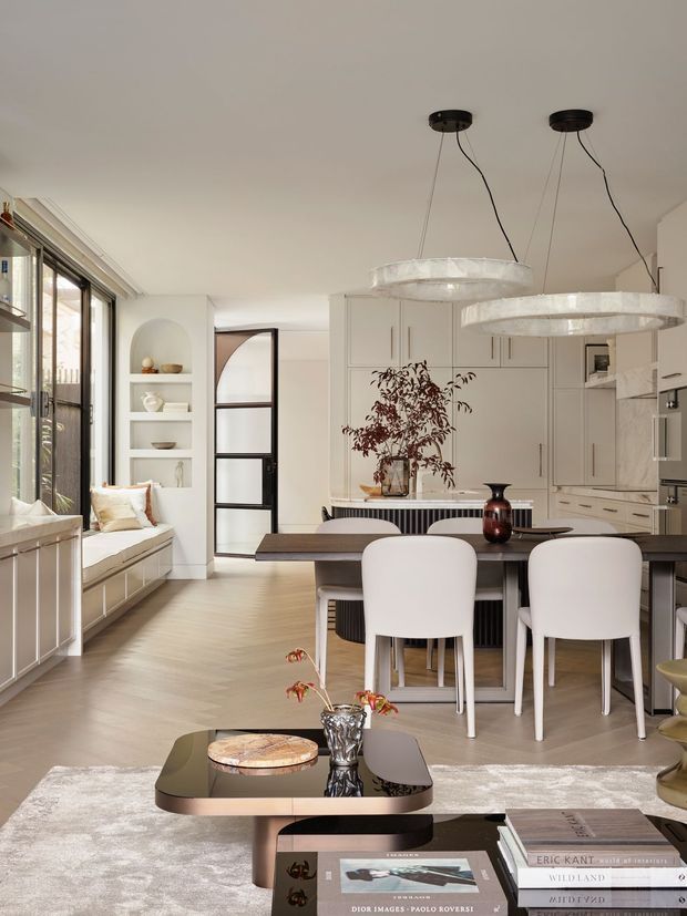 A bright, refined open-plan interior combining a living area, dining space and contemporary kitchen. The room features pale timber flooring, white built-in cabinetry, integrated shelving, a dark dining table with sculptural white chairs, a built-in window seat, large glazed openings and oversized translucent pendant lights. A glossy dark coffee table and neutral rug anchor the foreground.