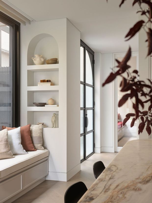 A bright, refined open-plan interior featuring a built-in window seat with neutral cushions, an arched display niche and open shelving, black-framed glazed doors, and a large marble-topped island or counter with black stools. Pale walls, light timber flooring, and warm neutral furnishings create a calm contemporary setting.