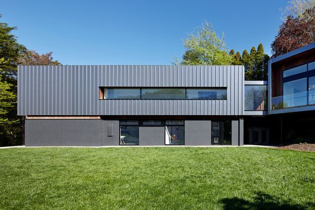 A contemporary two-storey residential building viewed from a broad lawn. The main rectangular volume has dark grey vertically ribbed metal cladding, a long horizontal strip window, dark lower-level walls and glazing, and a contrasting timber-toned connection to a projecting glazed wing on the right. Mature trees surround the property.