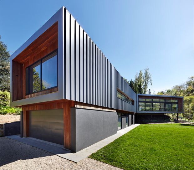 Modern two-storey residence with a sharply projecting linear form, dark vertical ribbed metal cladding, warm timber-lined wall sections, expansive glazing, and an integrated garage. The house sits beside a neatly landscaped lawn and wooded garden under clear blue sky.