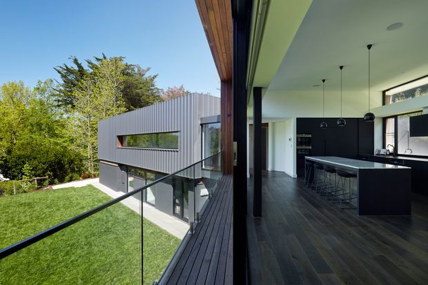 Split architectural view showing a contemporary home exterior and open-plan kitchen. The exterior features a dark grey corrugated metal-clad upper volume, extensive glazing, a glass balustrade, and a dark timber deck overlooking a landscaped lawn. The interior side shows a sleek black kitchen with a large island, dark flooring, white walls and ceiling, black pendant lights, and strong indoor-outdoor visual connection.