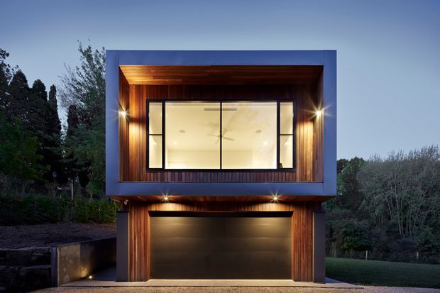 Night-time exterior view of a compact modern two-storey building, with a projecting upper volume clad in dark blue-grey panels and recessed warm timber-lined soffits and walls. A large illuminated window spans the upper level, while the ground floor contains a wide dark garage door framed by timber cladding. Integrated exterior lighting highlights the entry and facade, with landscaped vegetation surrounding the structure.