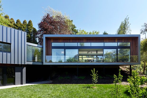 Contemporary elevated residential building with a rectilinear box form, extensive floor-to-ceiling glazing, and a warm timber-lined framed facade. The upper level projects over a dark recessed lower level, while a contrasting white and grey metal-clad wing connects on the left. Landscaped lawn and mature planting surround the structure.
