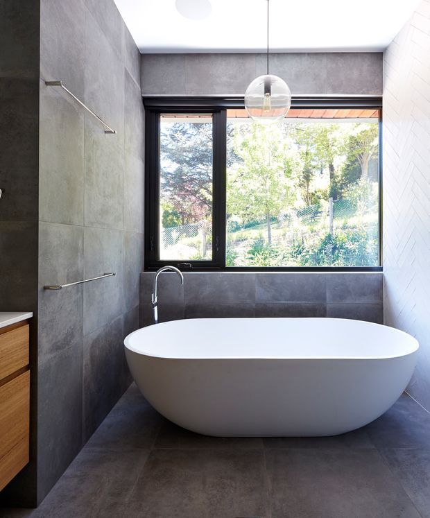 Contemporary bathroom featuring a freestanding white oval bathtub beneath a clear globe pendant light. Large dark grey tiled walls and floor create a cohesive, moody finish, with black-framed picture windows overlooking greenery. A partial timber vanity and towel rails are visible at the left.