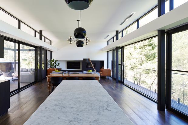 A bright, open-plan contemporary interior viewed from a large kitchen island, with dark timber flooring, extensive black-framed floor-to-ceiling glazing, a dining table, and a living area beyond. The space has a high ceiling with a partial mezzanine or upper-level void, sculptural black pendant lights, white walls, and strong views to surrounding trees through the glazing. A large pale stone island benchtop dominates the foreground.