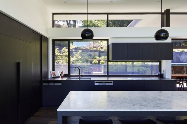Contemporary kitchen with dark, full-height cabinetry, a large pale stone island, black pendant lights, and expansive windows overlooking landscaped greenery. The kitchen has a clean-lined, high-contrast design with a partial dining area visible beyond.
