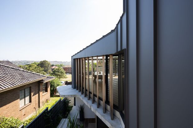 Exterior architectural view of a contemporary multi-level addition or residence, featuring a dark metal-clad facade, extensive floor-to-ceiling glazing with vertical timber or metal fins, and projecting concrete balconies. A neighbouring brick house and suburban landscape are visible beyond.