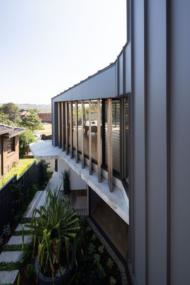 Exterior architectural view of a contemporary multi-level home on a sloped urban site. The image focuses on a dark grey metal-clad facade with tall glazing, vertical timber sun-shading fins, a projecting concrete balcony, and landscaped garden below. Neighbouring homes and a distant urban hillside are visible in the background.