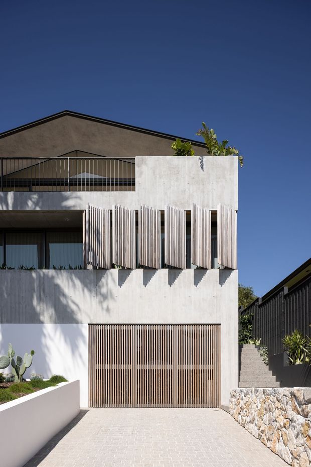 Street-facing contemporary multi-level residence with a pale concrete or rendered facade, strong vertical timber screening, a slatted garage door, recessed balcony, and rooftop planting. The home sits on a sloped site with a paved driveway, exposed stone retaining wall, and drought-tolerant landscaping under a clear blue sky.