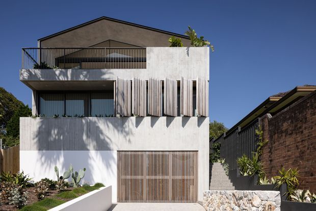 Street-facing view of a contemporary multi-storey residence with a white rendered and concrete facade, strong horizontal volumes, a dark pitched roof, recessed glazing, and prominent vertical timber or metal screening. The home includes a slatted garage door, rooftop terrace railing, landscaped entry planting, and a rough stone retaining wall.