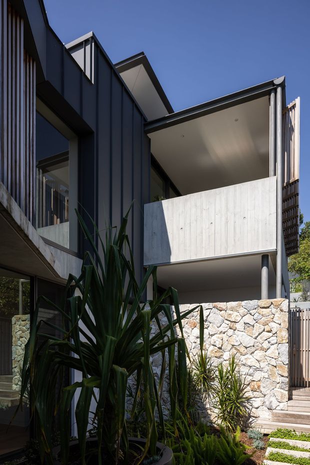 A close-up exterior view of a contemporary multi-level residence with dark vertical metal cladding, projecting flat-roofed balconies, exposed concrete surfaces, slender metal balustrades, and a substantial natural stone retaining wall. Lush architectural planting and stepping pavers frame the lower foreground.