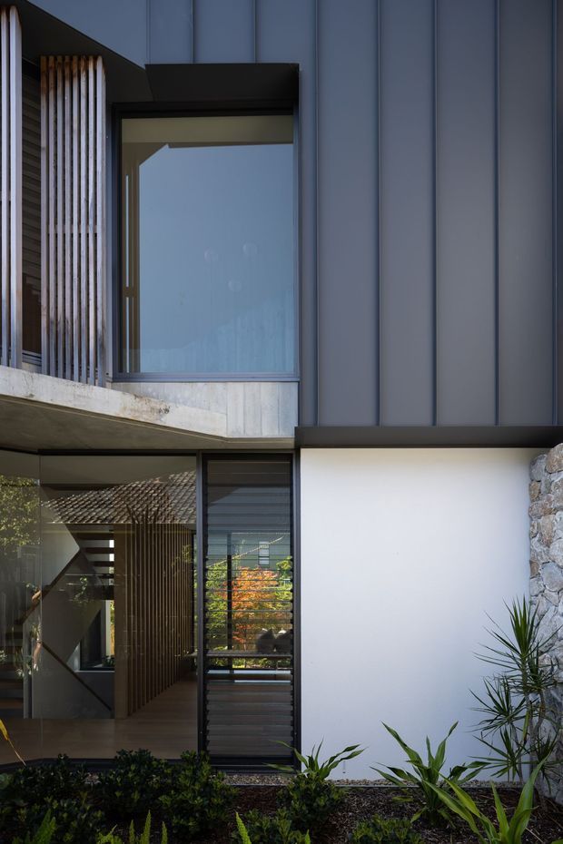 A close exterior view of a contemporary multi-level home facade. Dark vertical metal cladding and black-framed glazing contrast with a smooth white rendered wall, exposed concrete balcony edges, and a rough natural-stone feature wall. Vertical timber screening, a glazed entry, horizontal louvres, and landscaped planting are visible around the entrance.