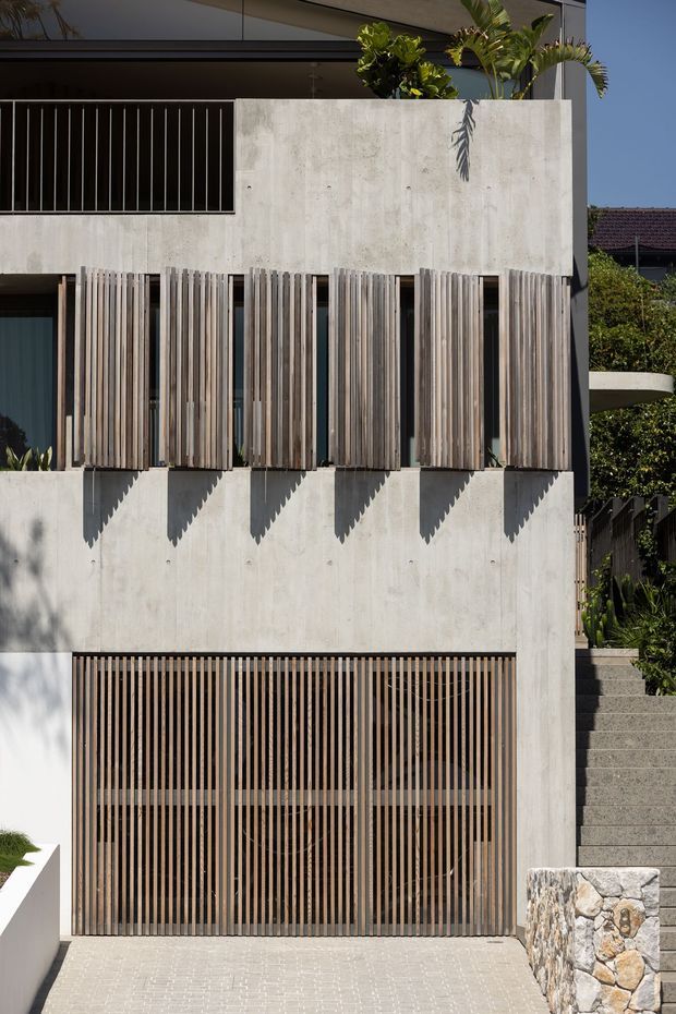 Vertical photograph of a contemporary multi-storey residential facade with exposed pale concrete walls, extensive vertical timber screening, dark metal balcony balustrades, and a slatted garage door. The facade is set on a sloping site with exterior steps, stone retaining walls, and planted balconies.