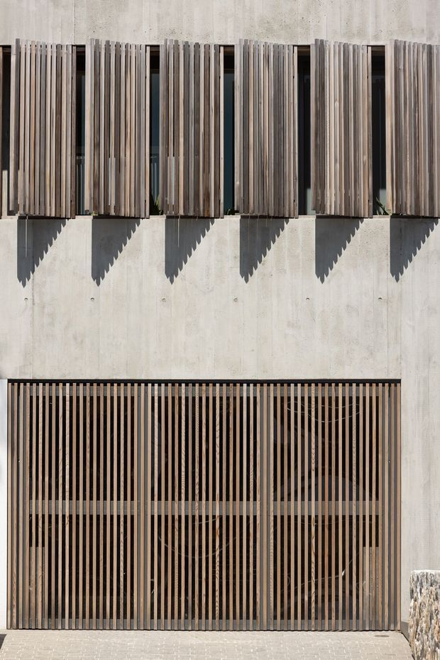 Close-up exterior view of a contemporary concrete building facade. Vertical timber and dark metal slatted screens shade a row of upper windows and a large lower glazed opening, creating strong repetitive lines and dramatic angular shadows in bright sunlight.