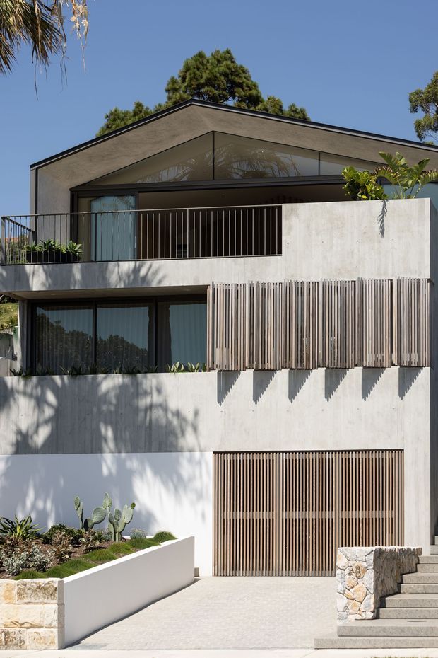 Modern multi-level residential facade with a flat roof, smooth light concrete or rendered walls, extensive dark-framed glazing, and vertical timber screening across balconies and garage doors. The house is set on a sloping site with landscaped planter beds, a paved driveway, and stone retaining elements. Strong geometric lines, deep overhangs, and integrated privacy screens define the exterior.
