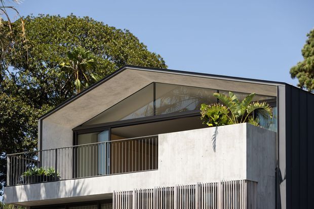 Contemporary two-storey residential facade with a sharply angled gable-like roofline, expansive glazing, dark metal cladding, and pale textured concrete walls. A projecting balcony with slim vertical metal balustrades and a raised planter adds greenery to the modern exterior.