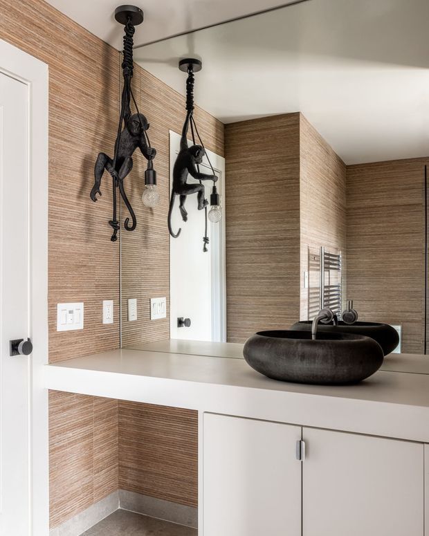 Contemporary bathroom vanity with a large wall mirror, white built-in cabinetry and benchtop, dark sculptural vessel basins, and a chrome heated towel rail. The walls feature warm horizontal wood-look textured panelling, complemented by playful black monkey pendant lights and dark hardware.