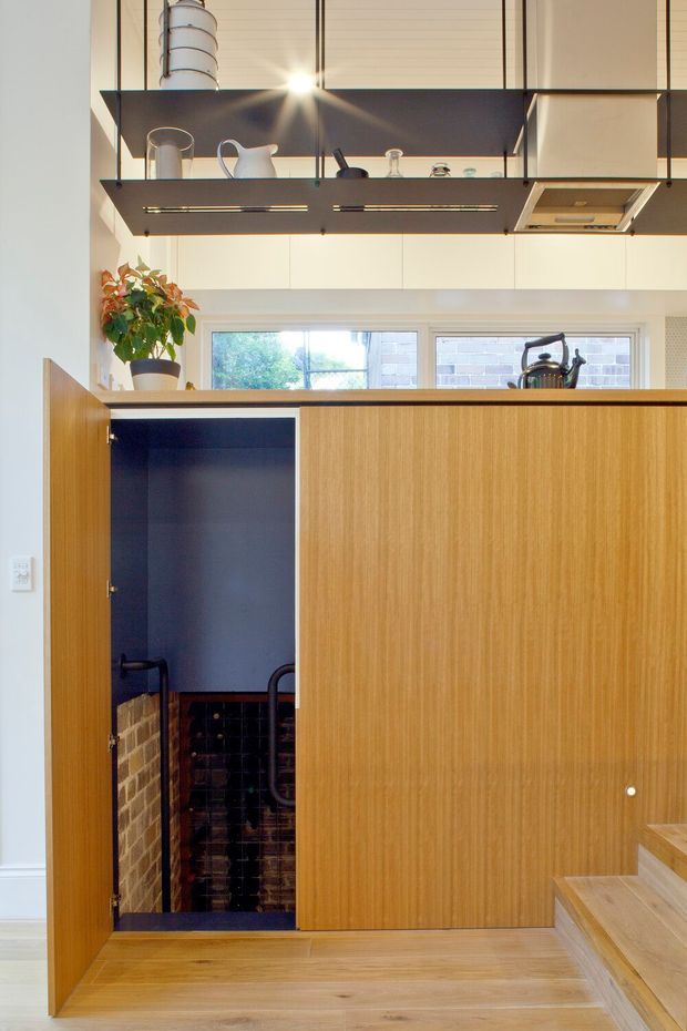 Bright contemporary interior featuring a compact kitchen and stair zone. A large vertical-grain timber-clad cabinet or partition dominates the foreground, with a concealed door open to a dark brick-lined storage area. Above, open dark shelving supports kitchenware and a cooker hood, while rear windows bring in daylight. Timber flooring and stairs continue through the space, creating a warm, clean, functional composition.