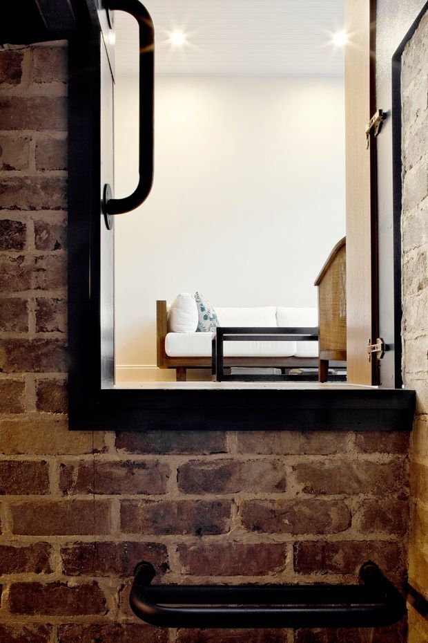 View through a black-framed internal window or pass-through in an exposed brick wall, looking into a bright minimalist living room. The room beyond contains a white sofa, dark rectangular coffee table, and warm timber cabinetry, while the foreground is dominated by rustic exposed brick, a bold black frame, and black hardware/handrail. Recessed ceiling lights and a restrained neutral palette create a strong industrial-contemporary contrast.