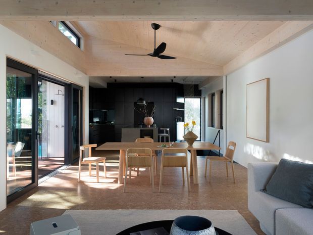 A bright, open-plan contemporary interior combining living, dining and kitchen areas. The room features exposed pale timber ceiling beams and matching timber wall and floor finishes, a matte-black kitchen with a central island, a timber dining table and chairs, large black-framed glazed doors, and a light upholstered sofa. Natural light creates a warm, airy atmosphere.