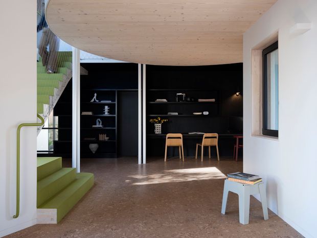 A contemporary open-plan interior featuring a sculptural curved timber ceiling, dark built-in shelving and joinery, a compact dining or study area, and a striking lime-green staircase with matching curved handrail. The space has polished brown-toned flooring, white walls, slim white structural columns, and strong natural light entering through the window and stairwell.