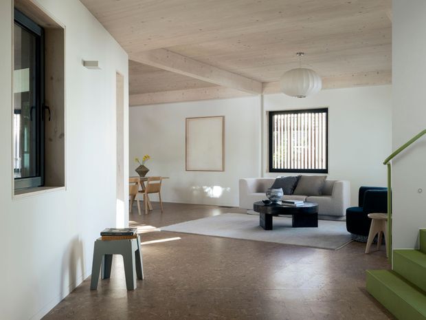 Bright, minimalist open-plan living room with pale timber-lined ceiling and exposed beams, white walls, polished concrete floor, and large dark-framed windows with vertical blinds. A light sofa, black circular coffee table, sculptural pendant, small dining area, and green-carpeted staircase create a restrained contemporary interior.