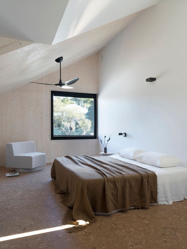 A serene contemporary bedroom with a low-profile bed dressed in white linen and a large taupe-brown throw, set against a white plaster wall and pale timber-panelled wall beneath an angular sloped ceiling. A black-framed window overlooks greenery, while a sculptural black ceiling fan, simple wall lights, and a pale upholstered lounge chair complete the minimalist interior. The room has a warm, natural material palette and abundant daylight.