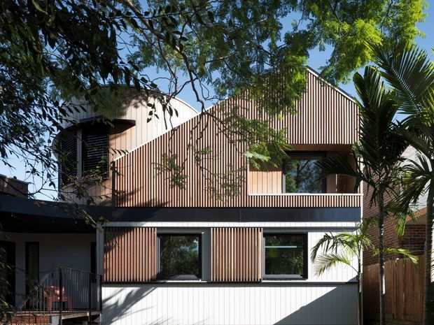 Exterior view of a contemporary multi-level residence with a distinctive angular pitched roof and vertically ribbed timber cladding. Dark horizontal trim separates the upper and lower levels, while large rectangular windows and a sheltered balcony or terrace add contrast. Mature trees and palms partially frame the facade.