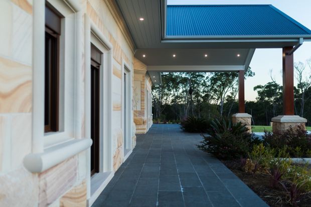 Exterior architectural view of a covered veranda or entrance walkway beside a sandstone-clad building. The facade has pale layered stonework with projecting trim around dark-framed windows and doors. A broad dark tiled or stone-paved path runs beneath a generous roof canopy supported by timber-toned square columns on stone bases. Recessed downlights illuminate the soffit, while landscaped planting and trees extend beyond the covered area. The composition emphasises the building facade, roof canopy, and outdoor circulation space.