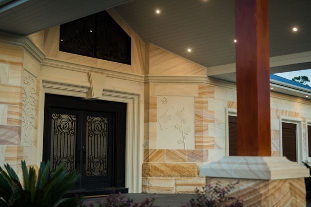A richly detailed covered entry porch with a black ornate double front door and matching decorative transom grille. The facade combines cream and warm sandstone-toned masonry with carved or etched decorative panels, classical mouldings, and a substantial timber support post. Recessed downlights illuminate the soffit, while planting and a low masonry balustrade frame the foreground.