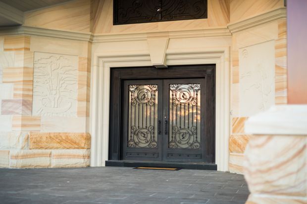 A formal building entrance viewed from outside, featuring ornate black double doors with decorative wrought-iron and patterned glass inserts. The doorway is framed by cream and pale stone cladding, carved relief panels, layered classical mouldings, and a dark slate-paved threshold. The composition has a grand, traditional character with warm neutral tones and strong black accents.