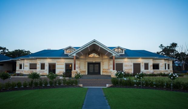 Front exterior view of a large, symmetrical single-storey residence with a broad blue pitched roof, prominent central gabled entry porch, cream rendered walls, stone-clad entry columns and facade accents, dormer windows, and dark-framed windows and doors. The building is approached by a straight paved path through a manicured lawn and formal garden.