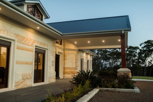 Exterior view of a substantial single-storey residence with a pale stone-clad facade, dark framed glazing, decorative gable detailing, and a broad covered entry or porte-cochère. The covered structure has a dark pitched metal roof supported by timber-toned posts with stone bases. Recessed downlights illuminate the entry, while landscaped garden beds and a gravel forecourt frame the approach.