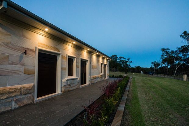 A single-storey contemporary building exterior photographed at dusk, with a long low facade clad in irregular light natural stone panels and dark-framed recessed doors and windows. Warm recessed eave lighting illuminates a paved walkway and narrow landscaped garden bed, while a broad lawn and trees extend alongside the building.