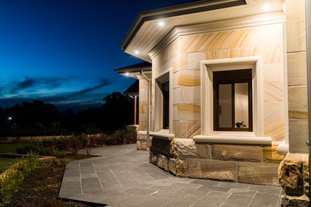 Night-time exterior view of a refined stone-clad building corner with recessed windows, heavy projecting trim, layered roof eaves, and warm architectural lighting. A paved walkway and landscaped planting border the facade, with a dark tree-lined setting beyond.